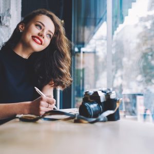 Positive female freelancer photography with red lips looking away and taking note while sitting at table with notebook and photo camera in modern workspace